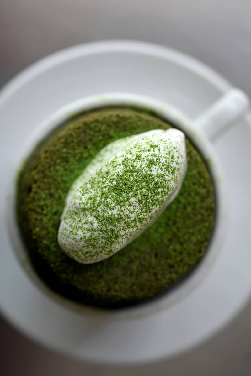 An overhead shot of white coffee mug filled with a gluten-free mug cake topped with coconut whipped cream and dusted with matcha powder. 